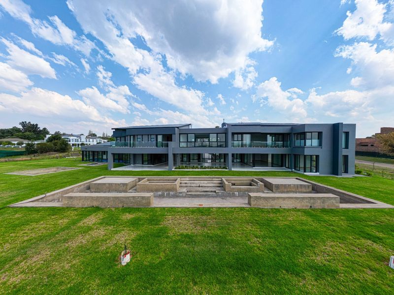 A modern, expansive rear elevation featuring large glass panels, multiple balconies, and a large lawn with an unfinished concrete water feature.