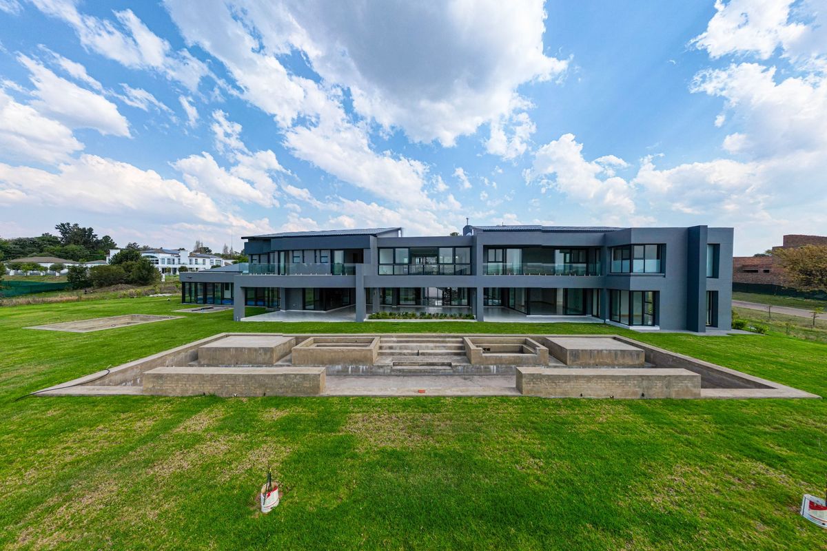 A modern, expansive rear elevation featuring large glass panels, multiple balconies, and a large lawn with an unfinished concrete water feature.