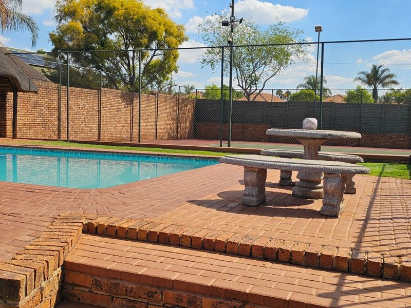Outdoor pool area featuring a swimming pool, thatched lapa, brick paving, and an adjacent tennis court with perimeter fencing.