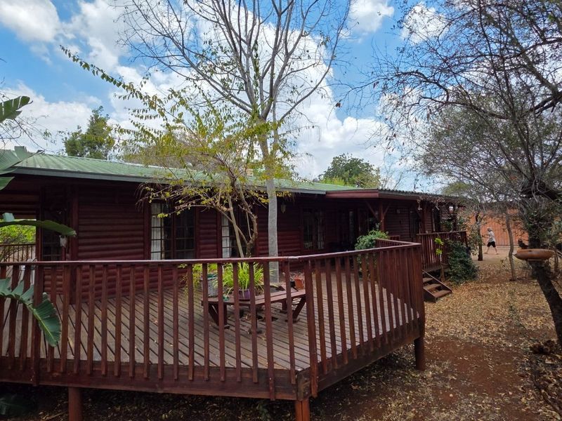 A rustic log cabin exterior featuring a wrap-around timber deck and balustrade, set within a natural, wooded farm landscape.