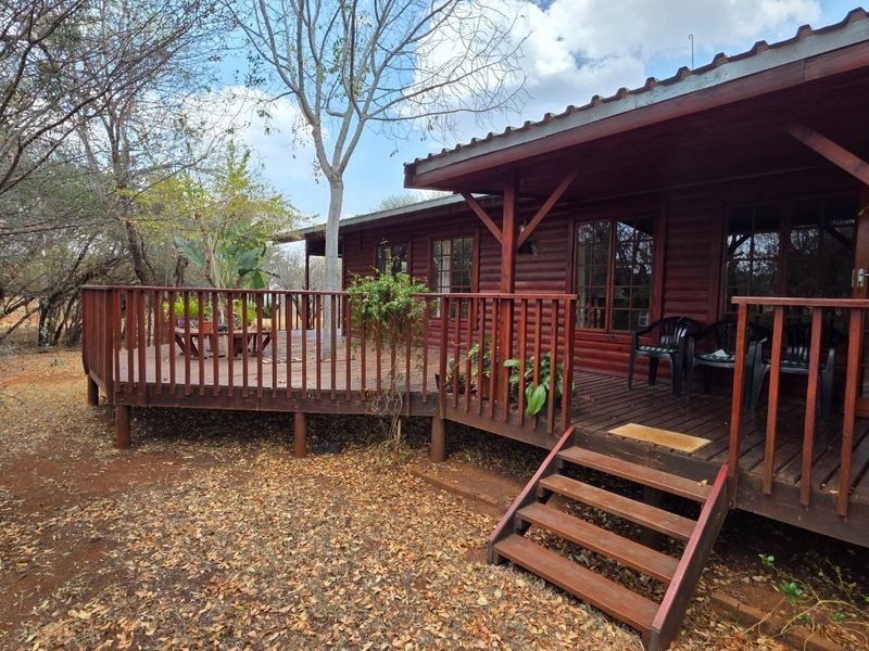 A rustic log cabin exterior featuring an expansive wrap-around timber deck, wooden balustrades, and a corrugated metal roof set within a natural bushveld environment.