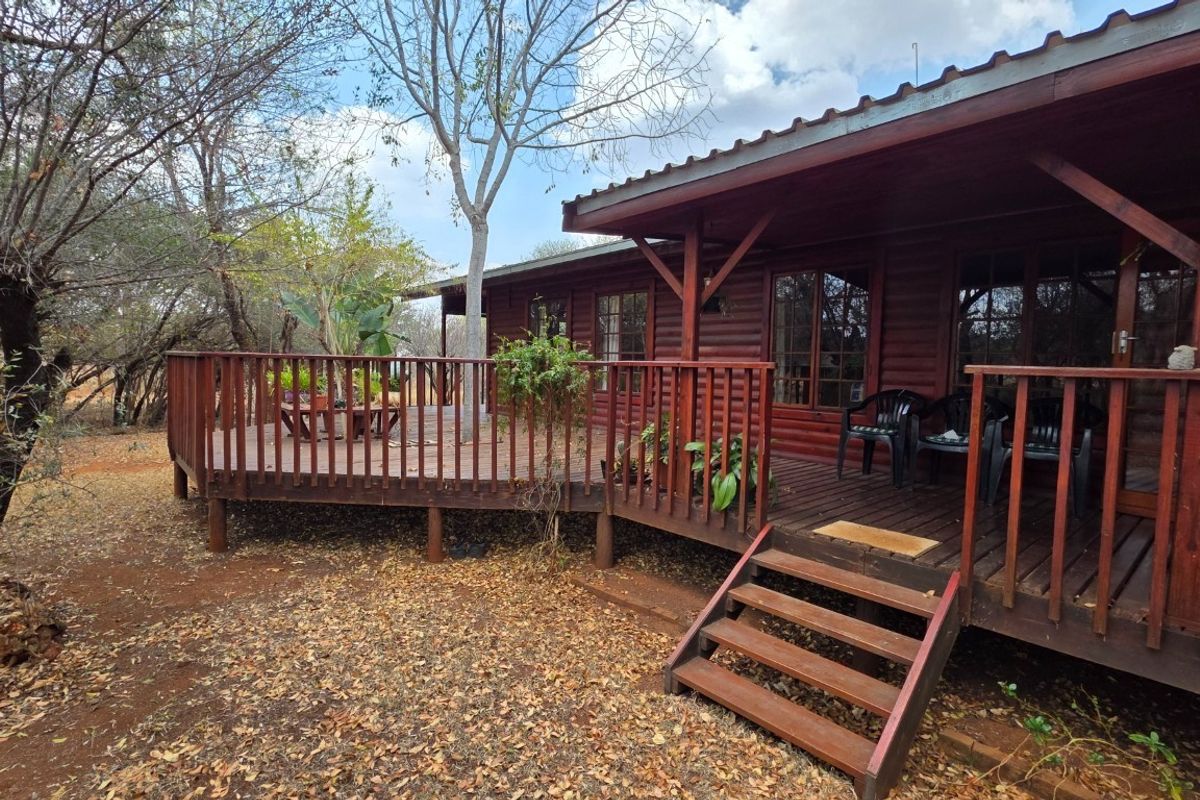 A rustic log cabin exterior featuring an expansive wrap-around timber deck, wooden balustrades, and a corrugated metal roof set within a natural bushveld environment.