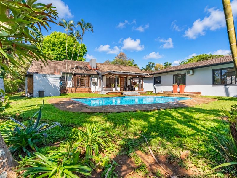 Rear view of the property featuring a swimming pool with stone surround, a covered patio area, and a spacious garden with established landscaping.