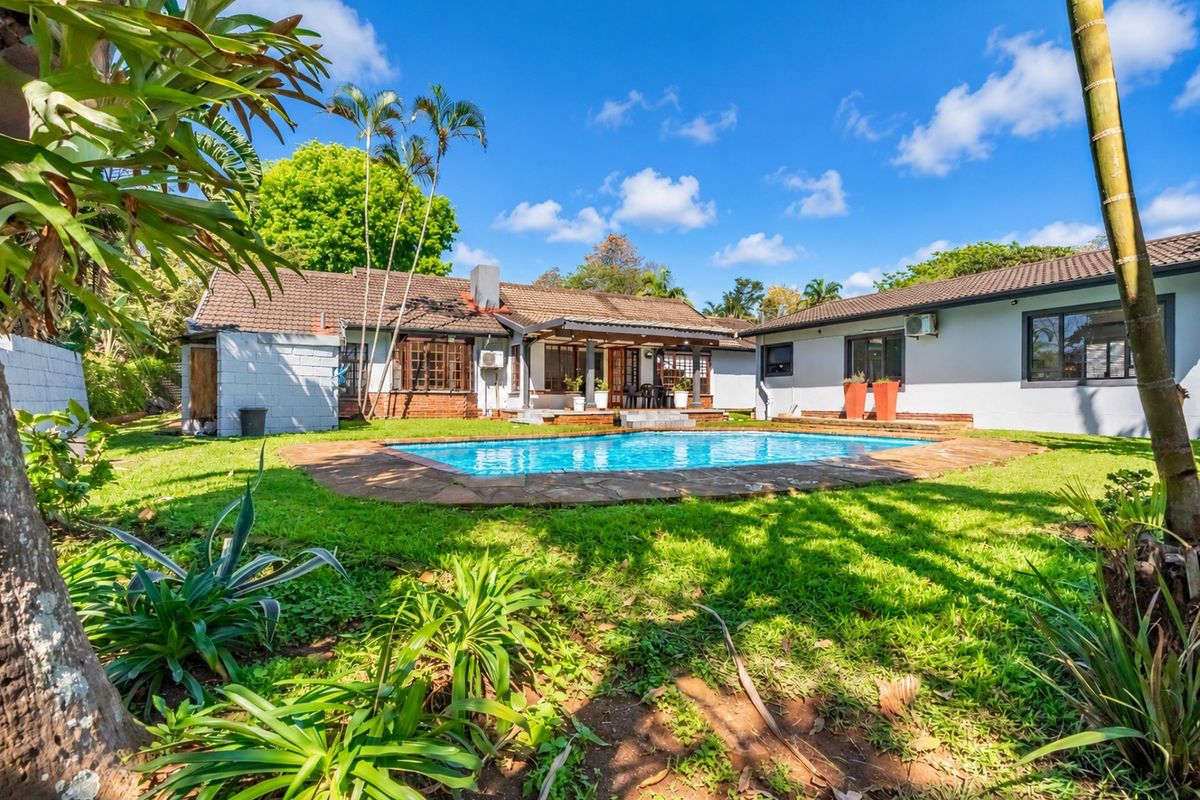 Rear view of the property featuring a swimming pool with stone surround, a covered patio area, and a spacious garden with established landscaping.