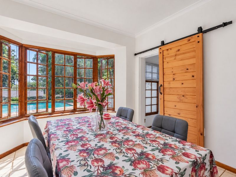 Bright dining room featuring a large bay window with garden and pool views, complemented by a rustic timber sliding barn door.