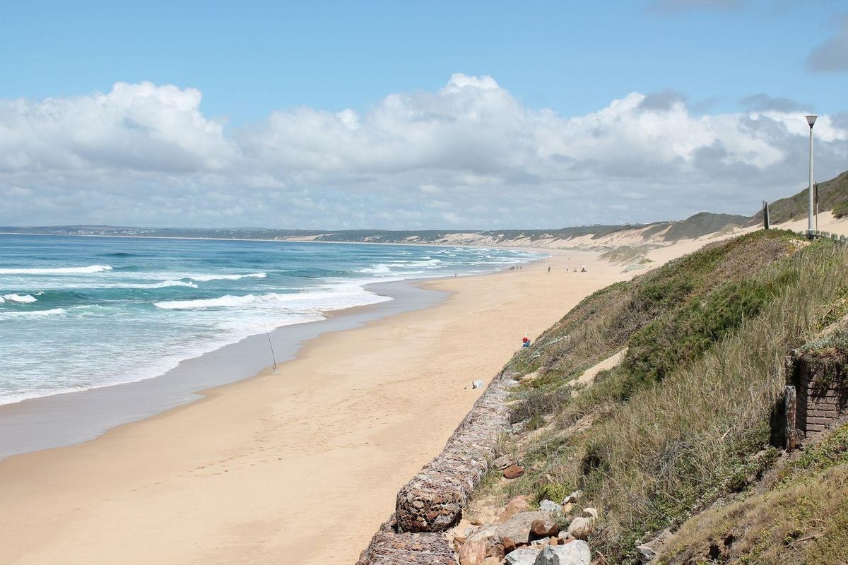 A scenic view of the expansive sandy coastline and ocean, featuring a protective gabion retaining wall along the elevated embankment.