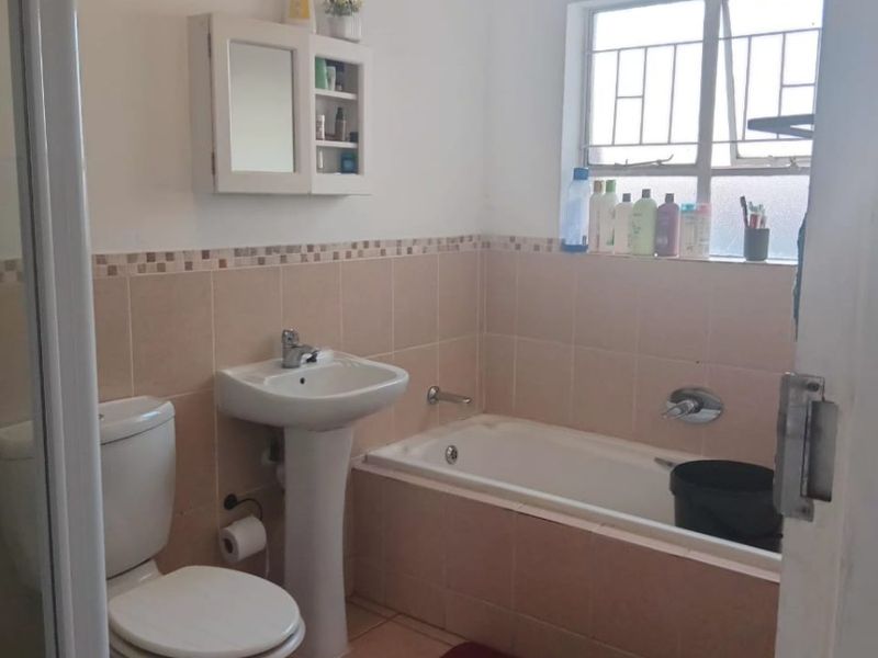 A functional bathroom featuring a pedestal basin, toilet, and bathtub with neutral wall tiling and a window with security bars.