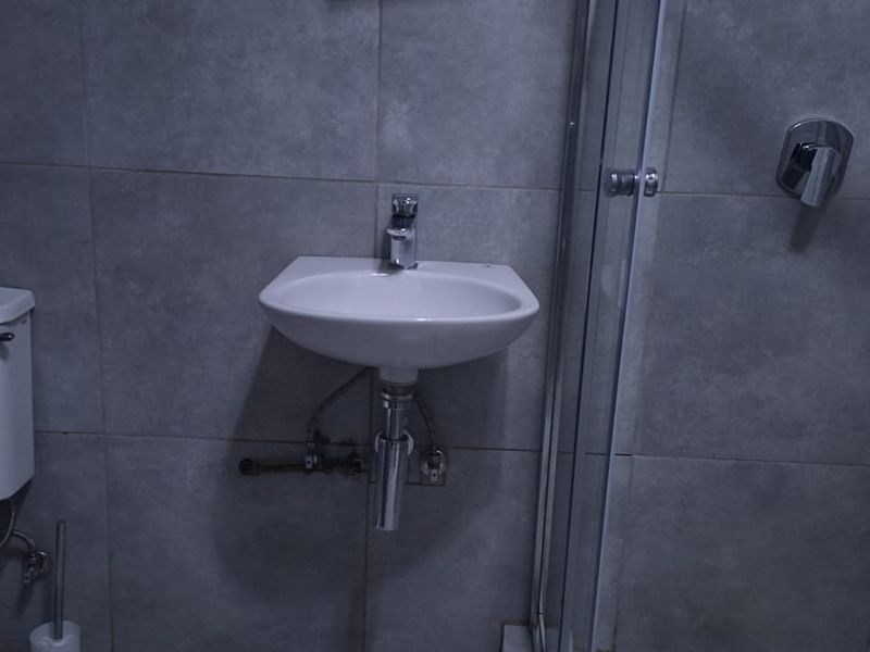 A compact bathroom featuring grey wall tiles, a wall-mounted basin, a toilet, and a glass-enclosed shower.