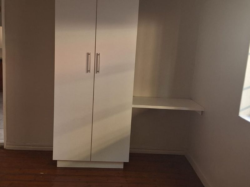 A bright, empty bedroom featuring timber flooring, a built-in wardrobe, and a small built-in desk area, with a view into an adjacent tiled room.