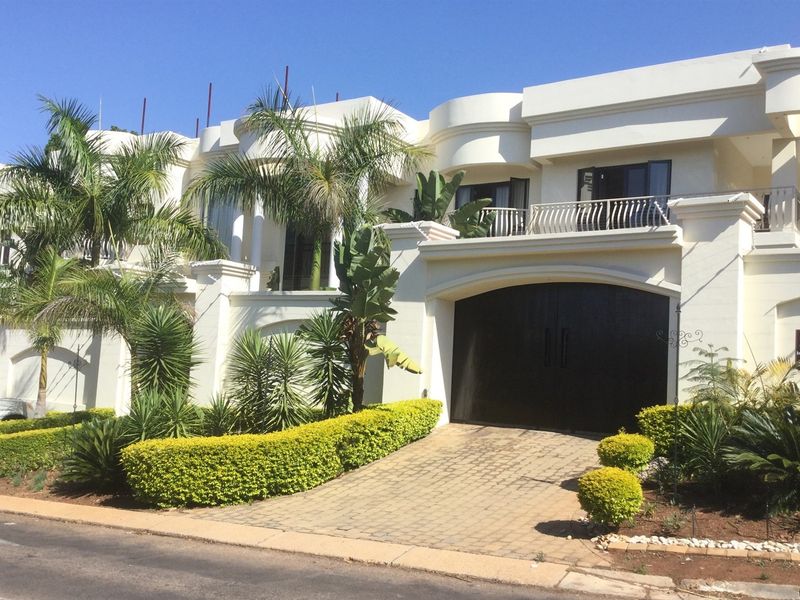 A modern multi-level residence featuring a paved driveway, double garage, and an upper-level balcony with metal railings, set within a landscaped garden.