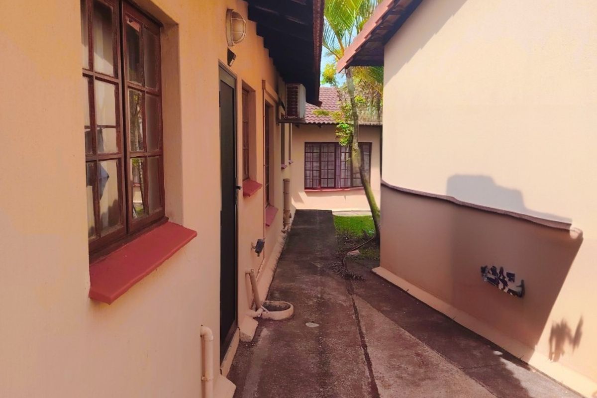 A narrow exterior side passage between two building sections featuring concrete paving, timber-framed windows, and an external air conditioning unit.
