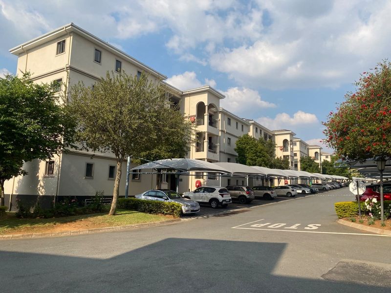 Exterior view of a multi-story apartment complex featuring paved driveways, covered parking bays, and well-maintained landscaped garden areas.