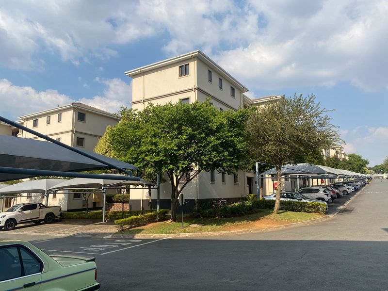 Exterior view of a multi-story apartment complex featuring paved driveways, covered shade cloth parking bays, and established garden landscaping.