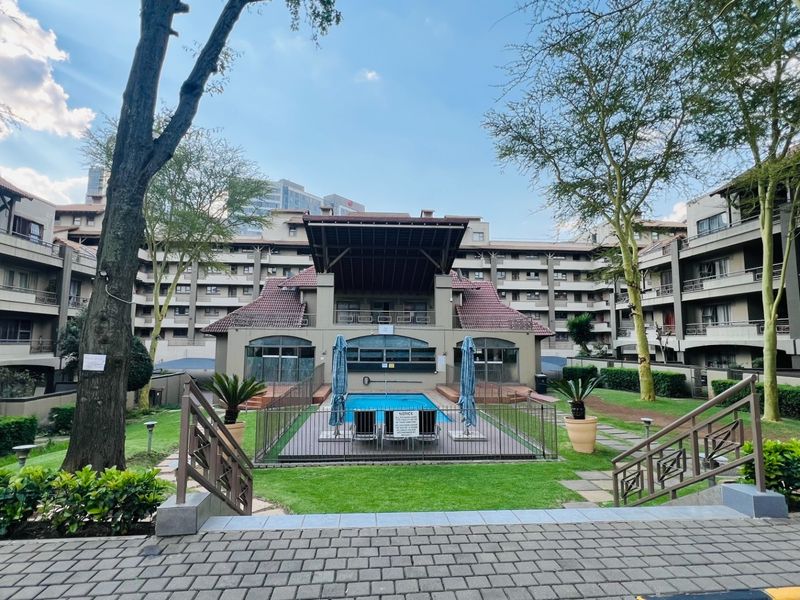 A well-maintained apartment complex courtyard featuring a central swimming pool, landscaped garden areas, and paved walkways surrounded by multi-story residential units.