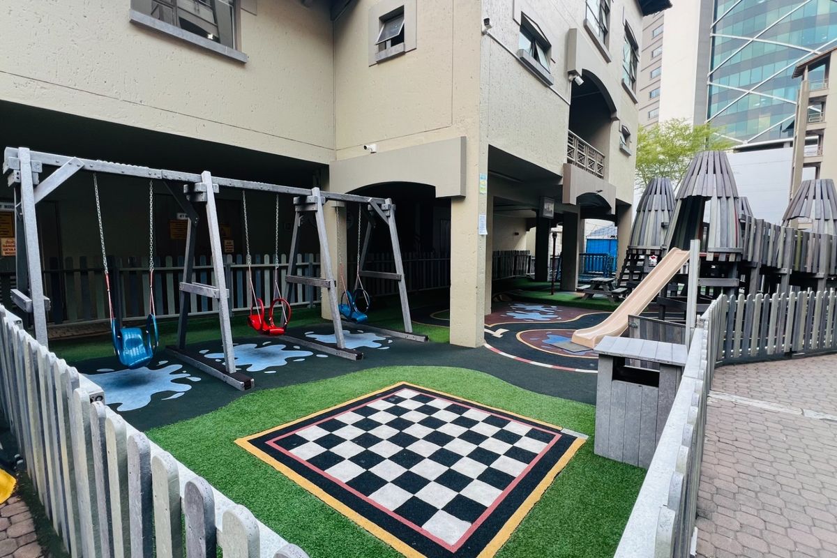 A communal outdoor playground area featuring synthetic grass, a built-in chess board, and various play structures within an apartment complex.