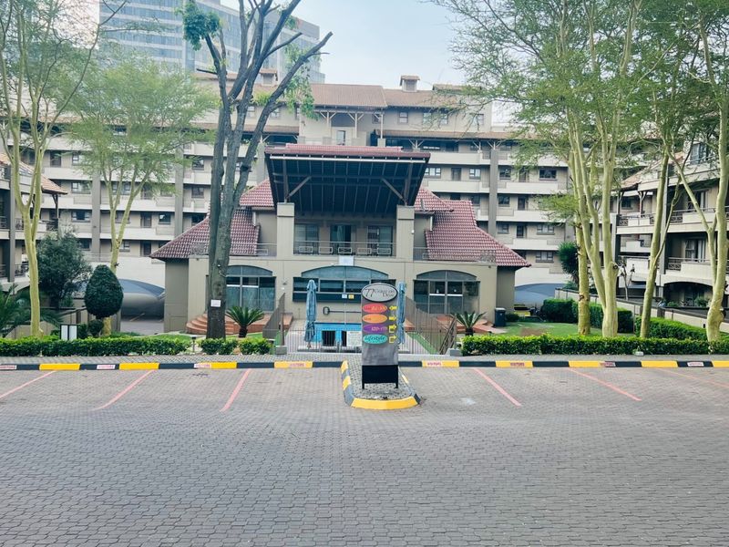 Front view of a multi-story apartment complex featuring a paved visitor parking area and established landscaped gardens.