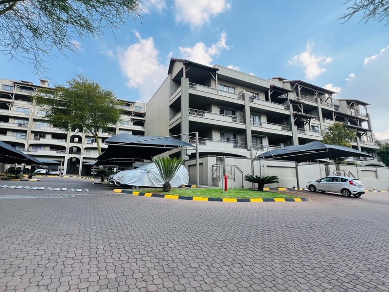 Exterior view of a multi-story apartment complex featuring paved parking areas, covered carports, and private balconies for each unit.