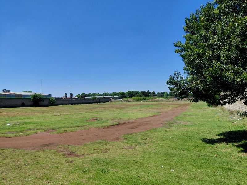A wide, open vacant commercial land plot featuring a grass lawn, dirt access track, and perimeter boundary walls.
