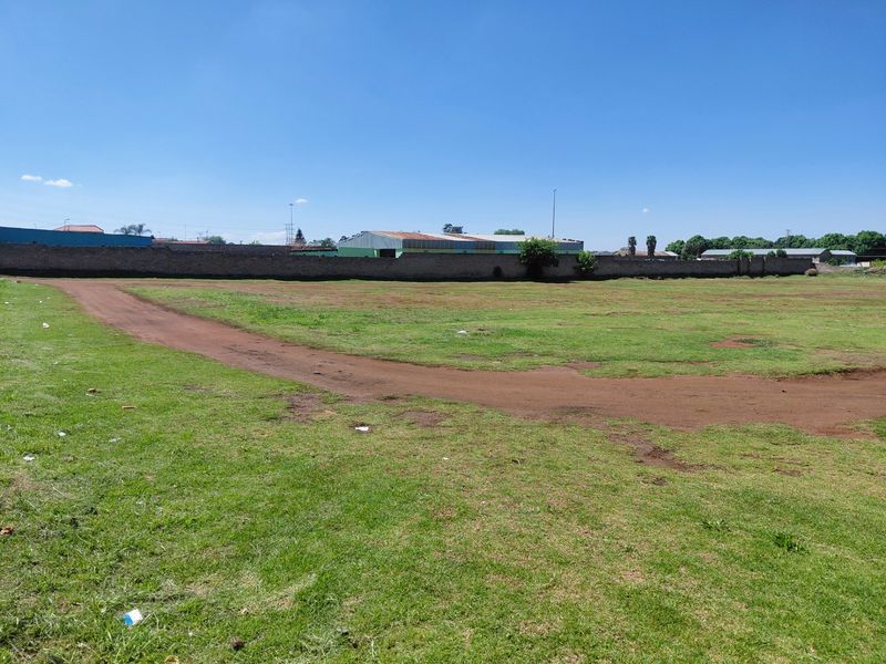 A wide, open commercial vacant land plot featuring a grass lawn and a perimeter boundary wall.