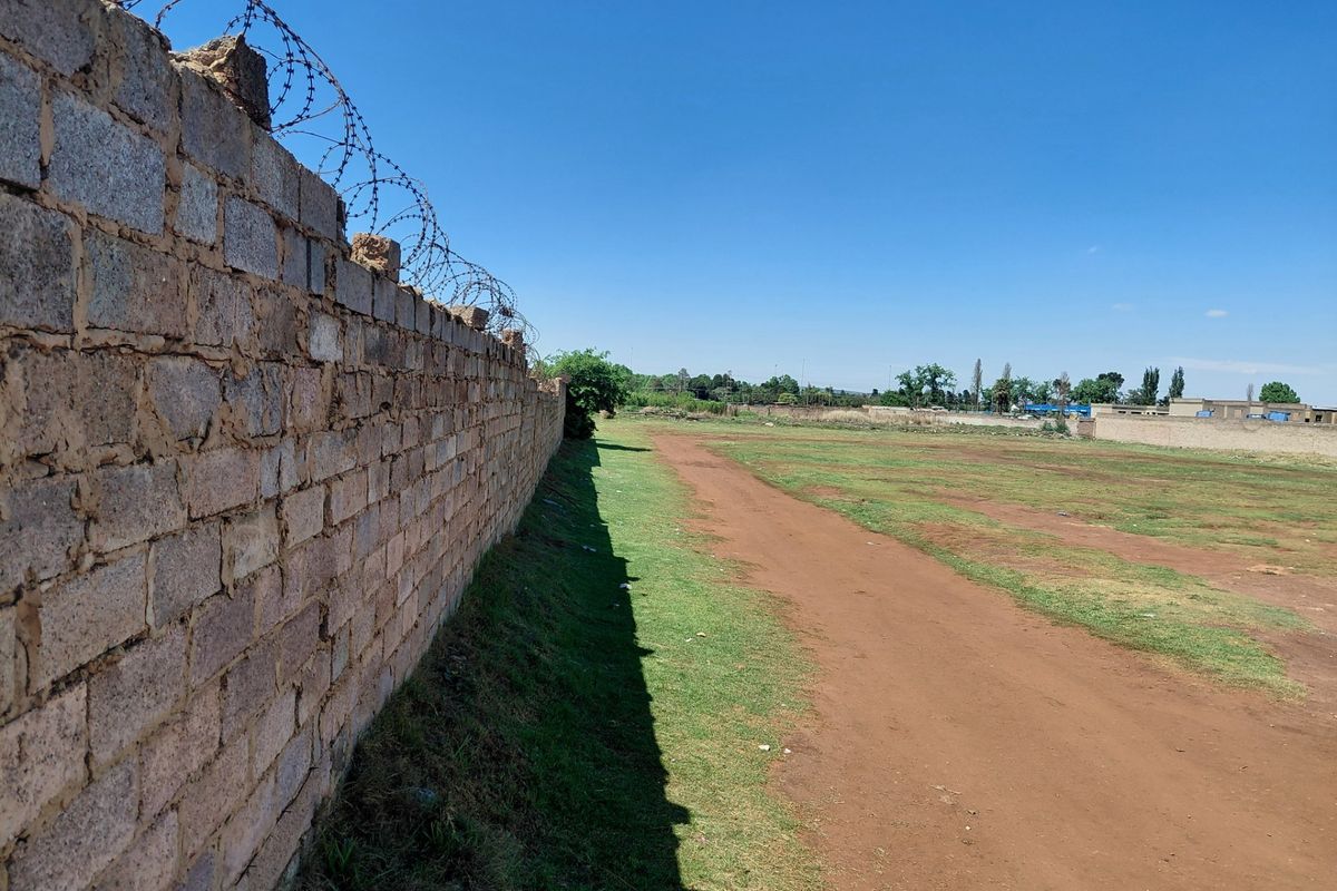 A view of a vacant commercial plot featuring a long perimeter brick wall topped with razor wire fencing and an open grass area.