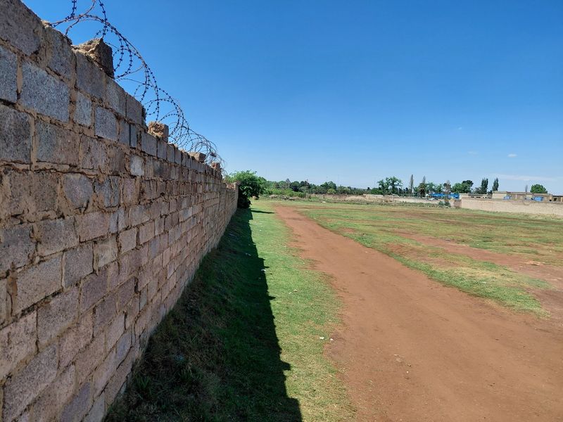 A view of a vacant commercial plot featuring a long perimeter brick wall topped with razor wire fencing and an open grass area.