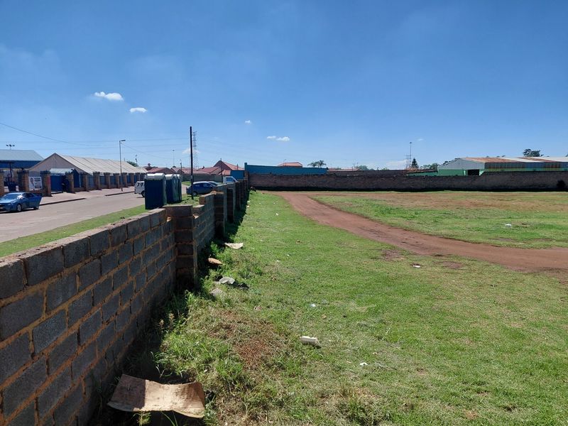 A vacant commercial land plot featuring a perimeter brick wall and grass lawn, situated along a street with surrounding industrial or commercial buildings.