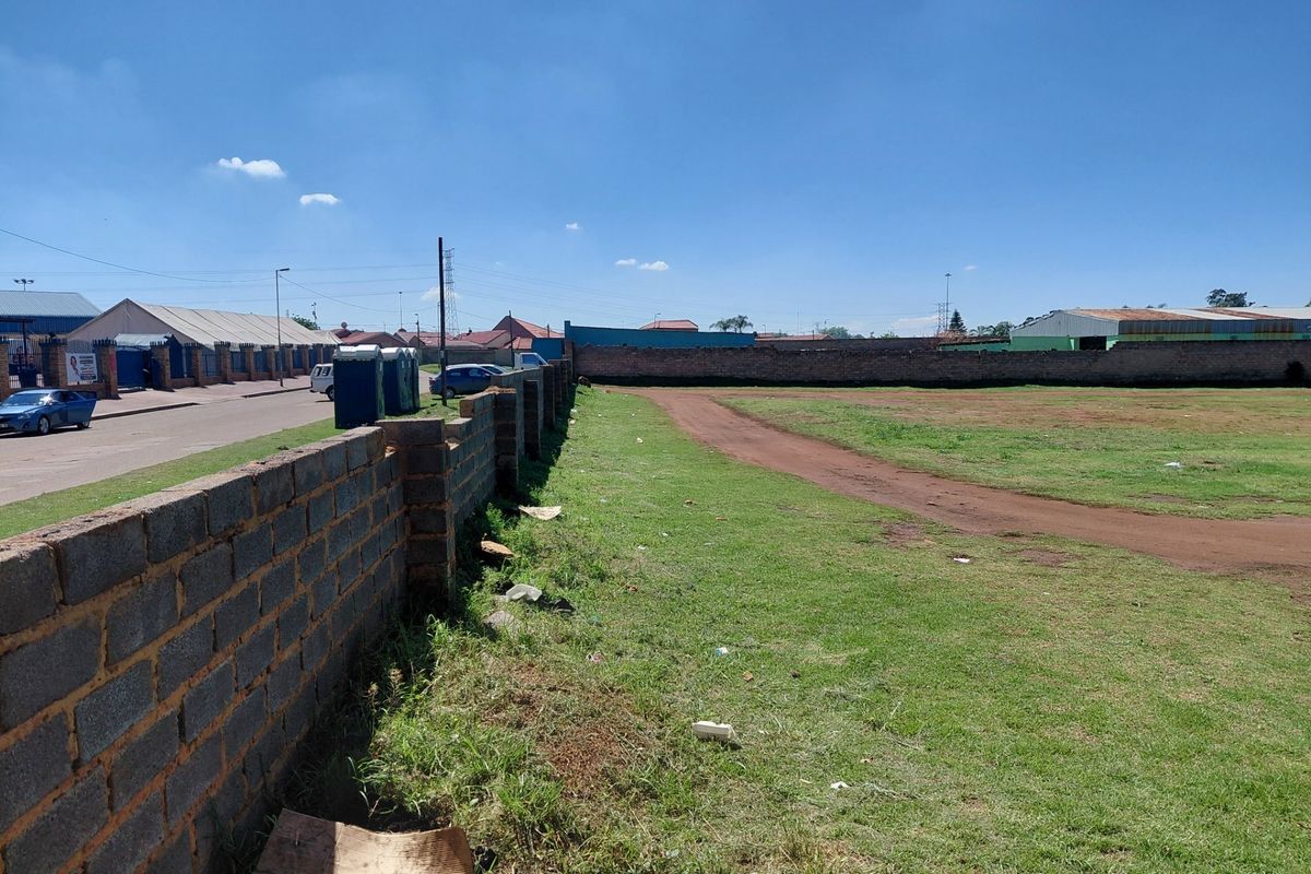 A vacant commercial land plot featuring a perimeter brick wall and grass lawn, situated along a street with surrounding industrial or commercial buildings.