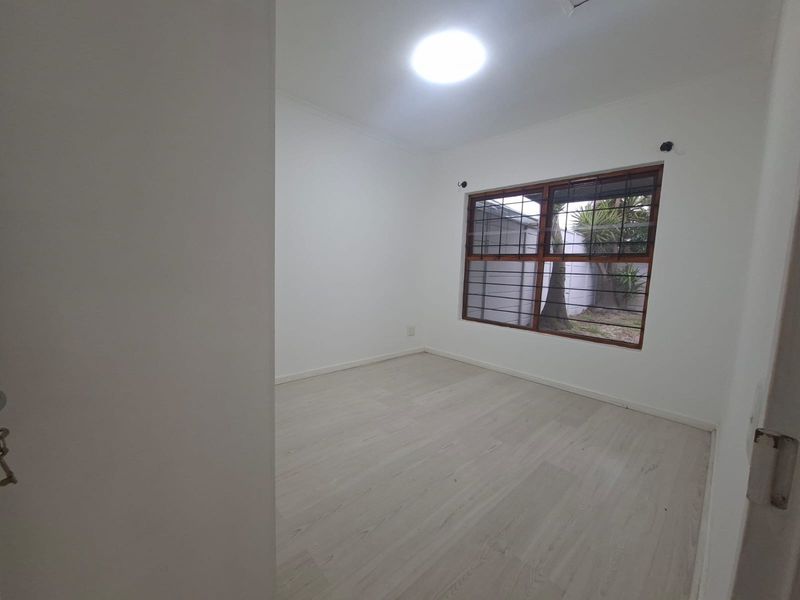 A bright, empty bedroom featuring light-toned laminate flooring, white walls, and a window fitted with security bars.
