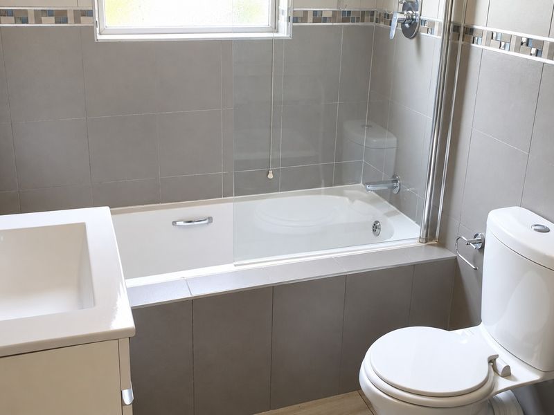A clean, modern bathroom featuring a built-in bathtub with a glass screen, a wall-mounted basin, and a toilet, finished with neutral grey wall tiles.