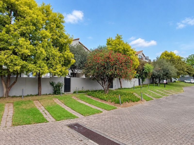 A well-maintained street view showing a paved driveway, perimeter wall, and landscaped verge with mature trees in a residential apartment complex setting.