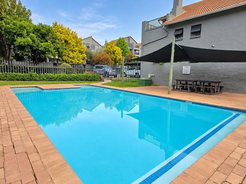 A well-maintained communal swimming pool area featuring a paved deck, shade sail, and surrounding landscaped lawn within a secure apartment complex.