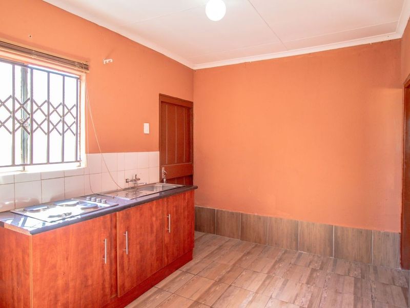 A compact kitchen featuring built-in wooden cabinetry, a stainless steel sink, a built-in hob, and tiled flooring with security bars on the window.