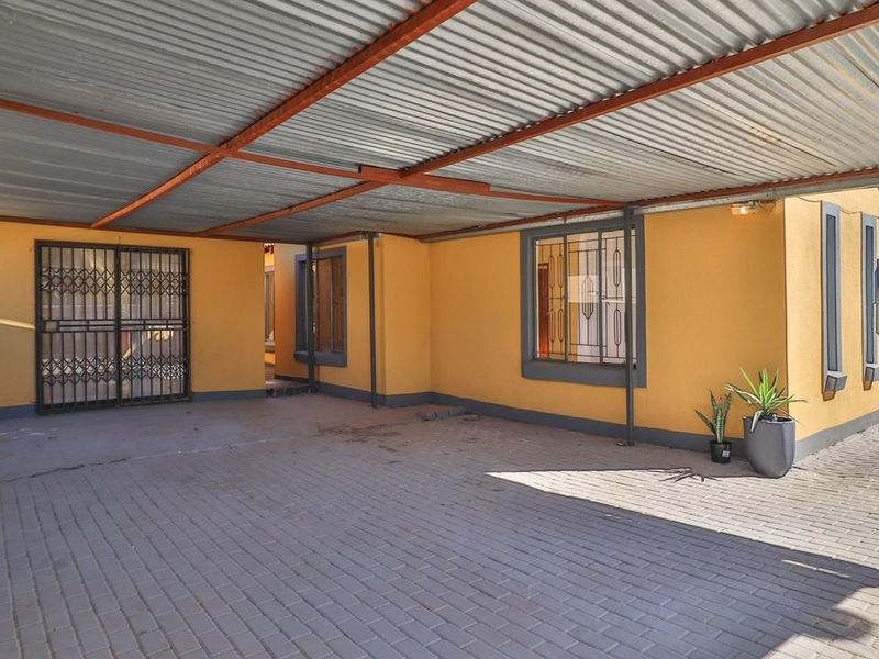 A covered carport area featuring a corrugated metal roof, paved flooring, and secure metal gates and window bars.