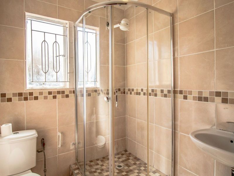 A functional bathroom featuring a corner shower enclosure, pedestal basin, toilet, and neutral ceramic wall tiling with a decorative mosaic border.