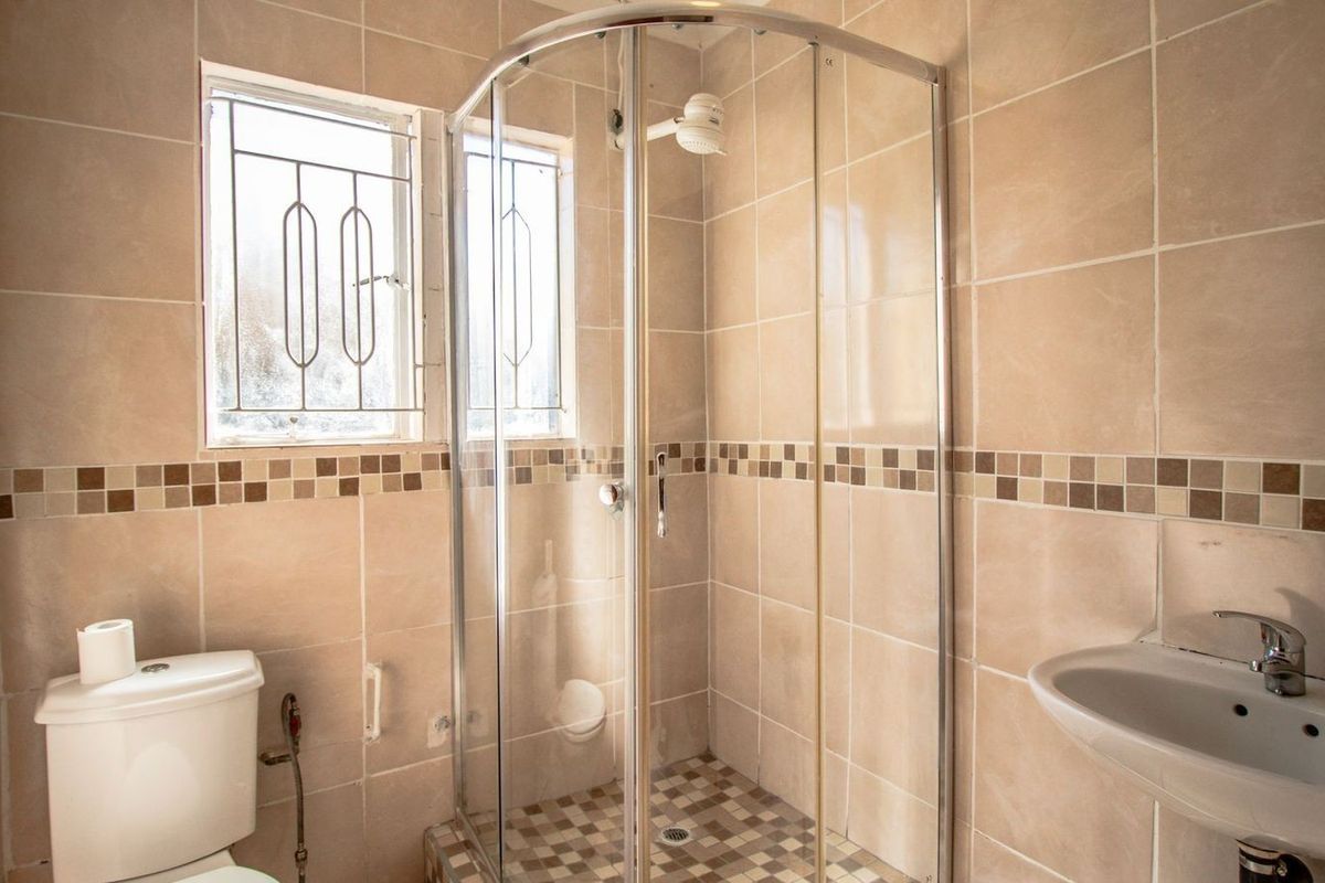 A functional bathroom featuring a corner shower enclosure, pedestal basin, toilet, and neutral ceramic wall tiling with a decorative mosaic border.
