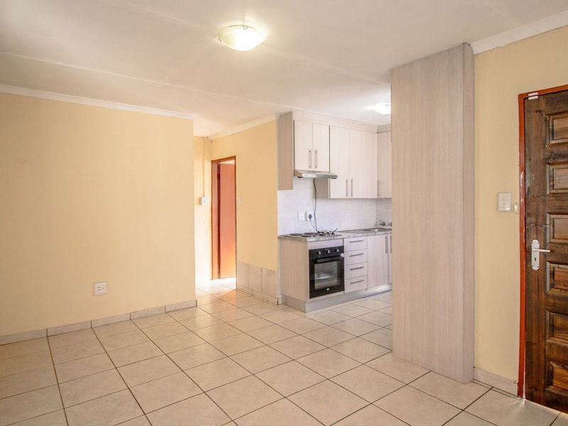 An open-plan living area featuring tiled flooring and a compact kitchen with built-in cabinetry, oven, and hob, leading to an adjacent room.