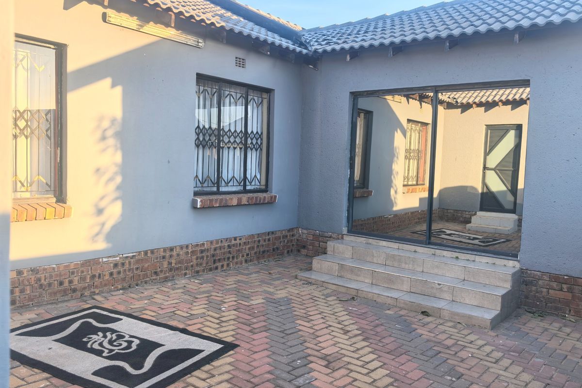 A rear exterior view of the house featuring a paved patio area, sliding glass door access, and security bars on the windows.