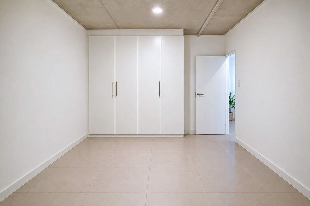 A clean, modern bedroom featuring neutral tiled flooring, built-in white cupboards, and an exposed concrete ceiling, offering a bright and minimalist living space.