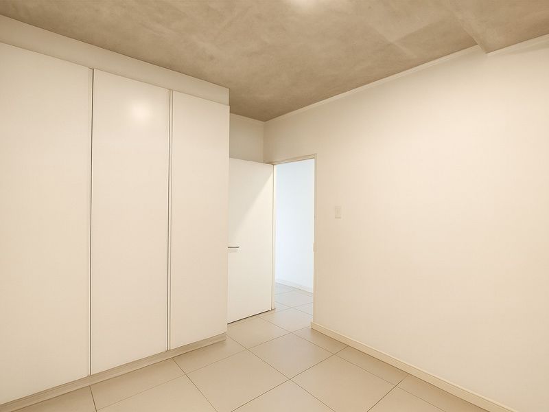 A clean, minimalist bedroom featuring built-in white wardrobes, neutral tiled flooring, and a raw concrete ceiling finish.