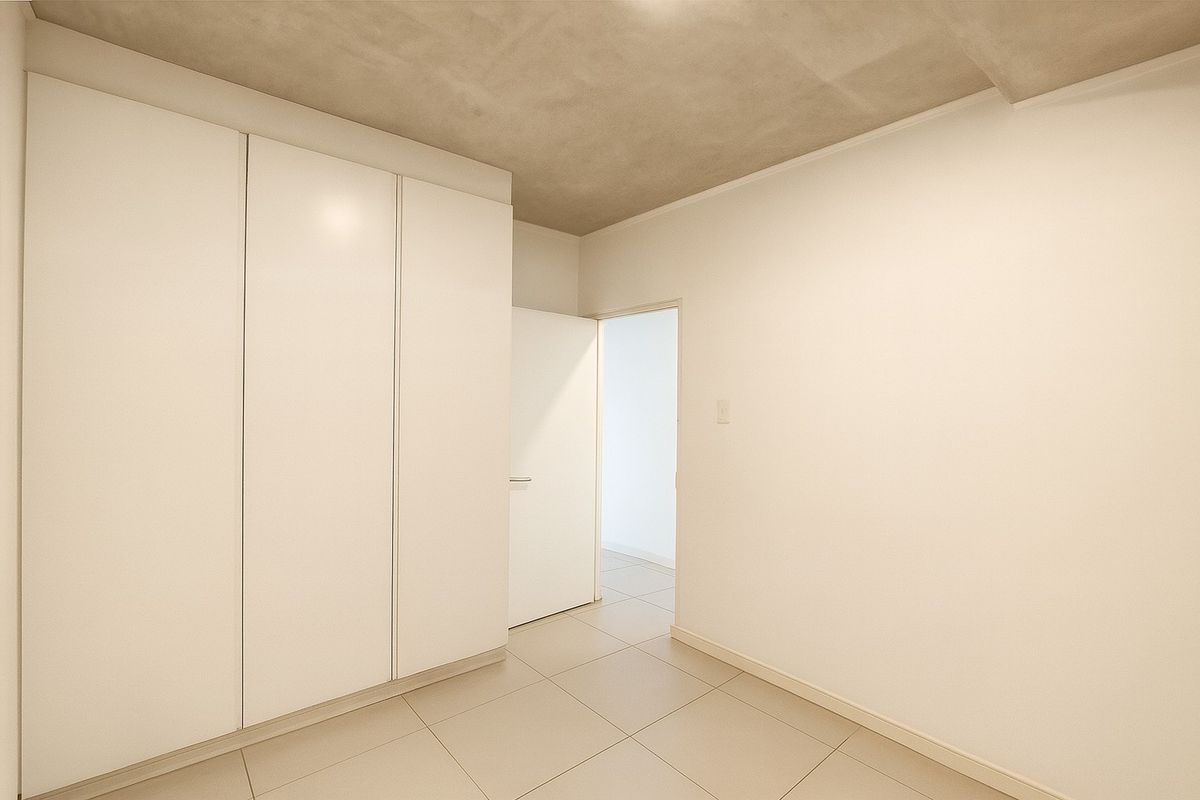 A clean, minimalist bedroom featuring built-in white wardrobes, neutral tiled flooring, and a raw concrete ceiling finish.