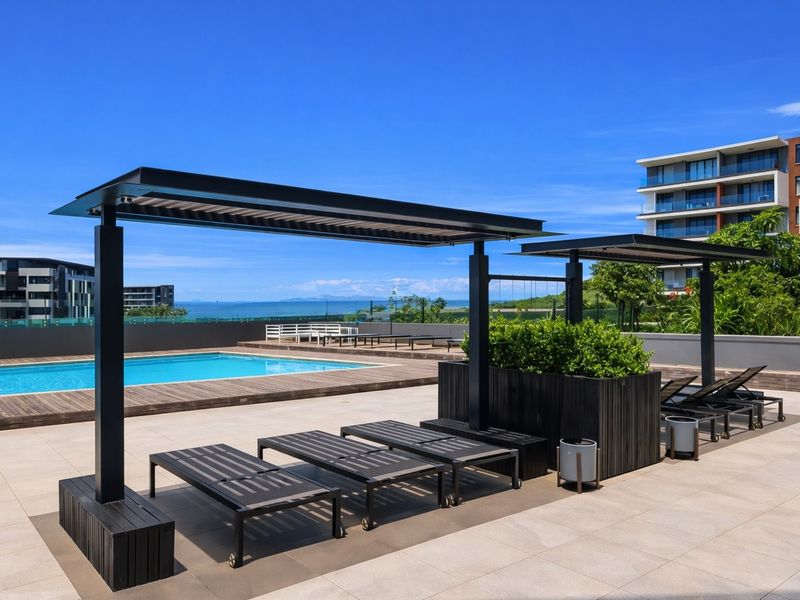 A well-maintained communal pool area featuring modern metal pergolas, timber decking, and a clear view of the ocean.