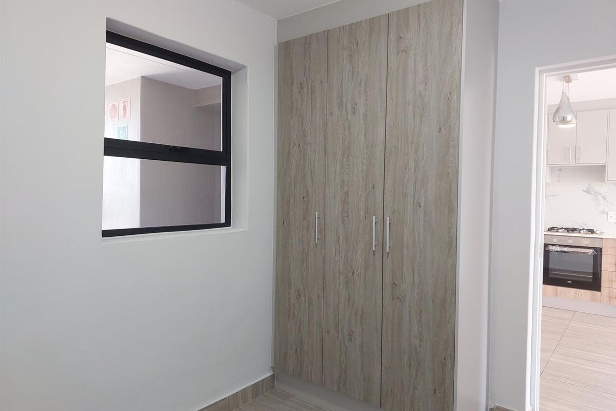 A clean, modern bedroom featuring built-in wood-look cupboards, tiled flooring, and a large window, with a view into the adjacent kitchen area.