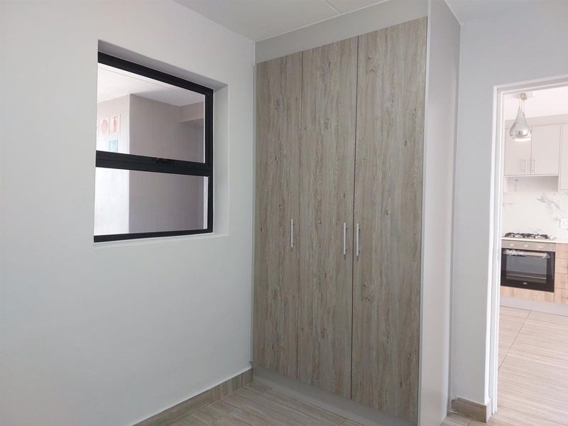 A clean, modern bedroom featuring built-in wood-look cupboards, tiled flooring, and a large window, with a view into the adjacent kitchen area.