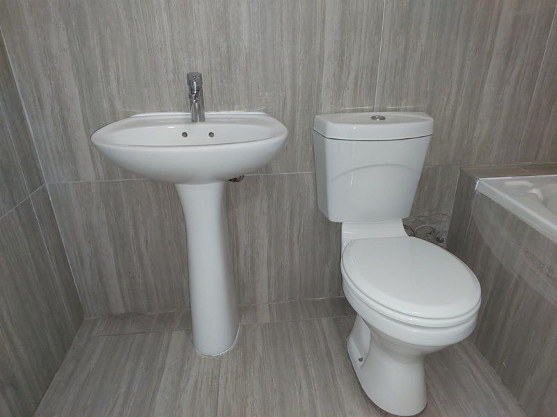 A functional bathroom featuring a pedestal basin, toilet, and bathtub with neutral wood-look wall and floor tiling.