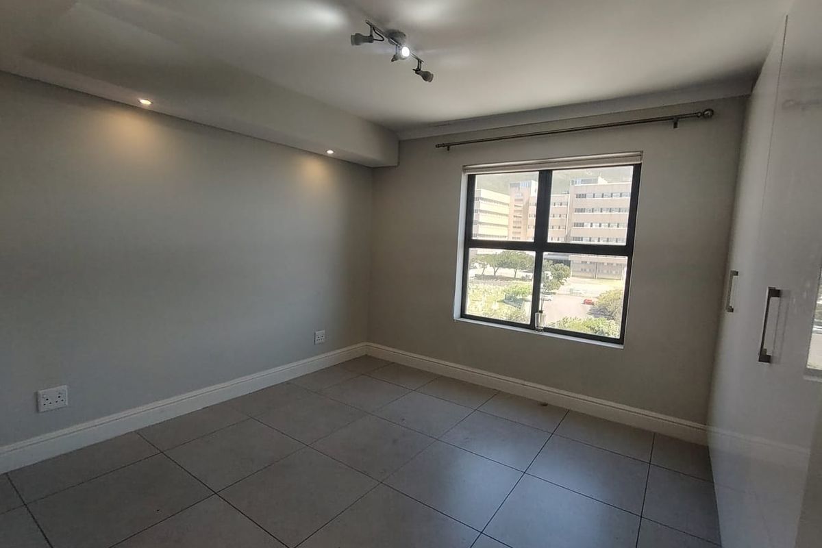 A bright, empty bedroom featuring neutral tiled flooring, built-in cupboards, and modern recessed and track lighting with a large window.