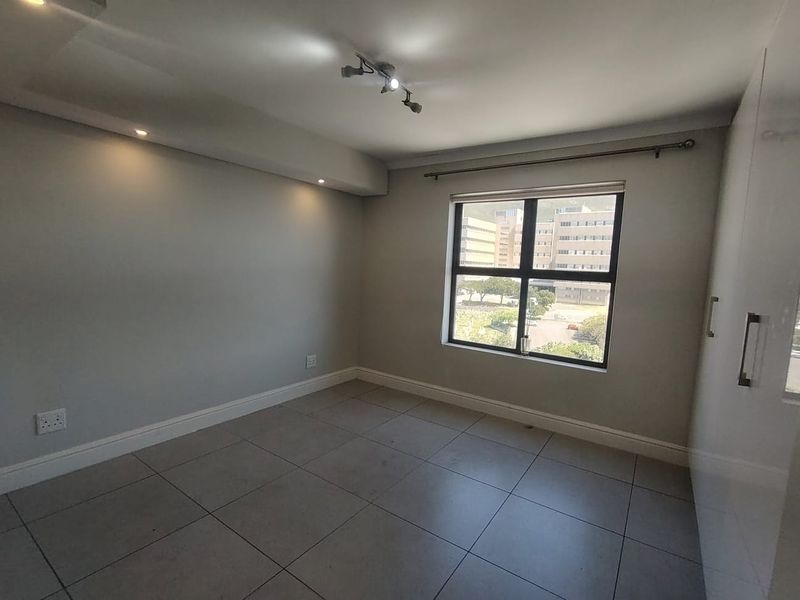 A bright, empty bedroom featuring neutral tiled flooring, built-in cupboards, and modern recessed and track lighting with a large window.