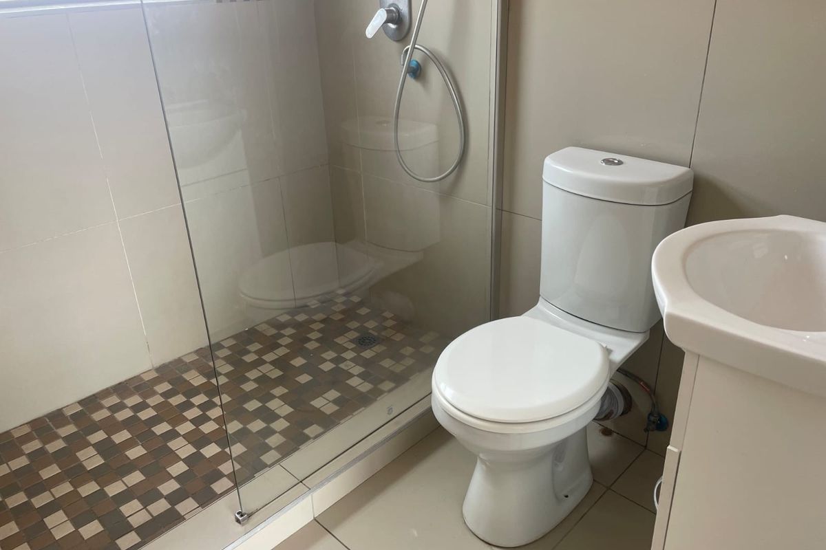 A functional bathroom featuring a glass-enclosed shower with mosaic tiling, a standard toilet, and a pedestal basin with neutral wall tiles.