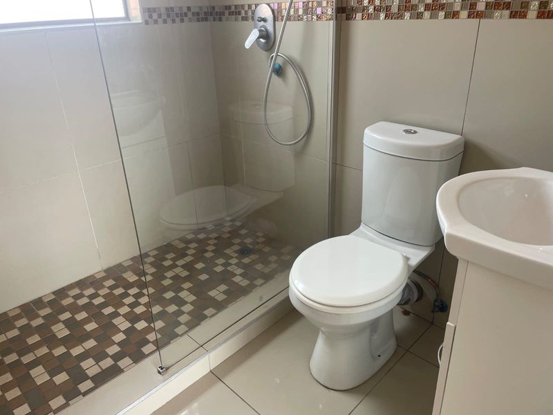 A functional bathroom featuring a glass-enclosed shower with mosaic tiling, a standard toilet, and a pedestal basin with neutral wall tiles.