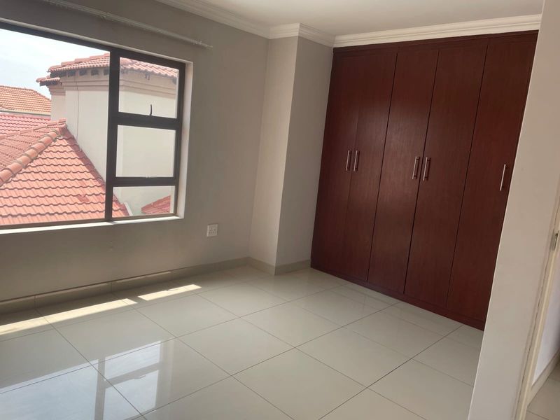 A bright, empty bedroom featuring neutral tiled flooring, built-in wooden cupboards, and a large window with views of the surrounding roofline.
