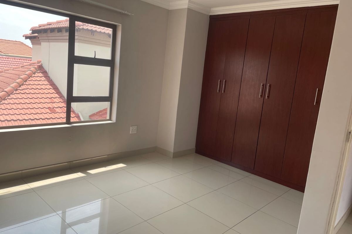 A bright, empty bedroom featuring neutral tiled flooring, built-in wooden cupboards, and a large window with views of the surrounding roofline.